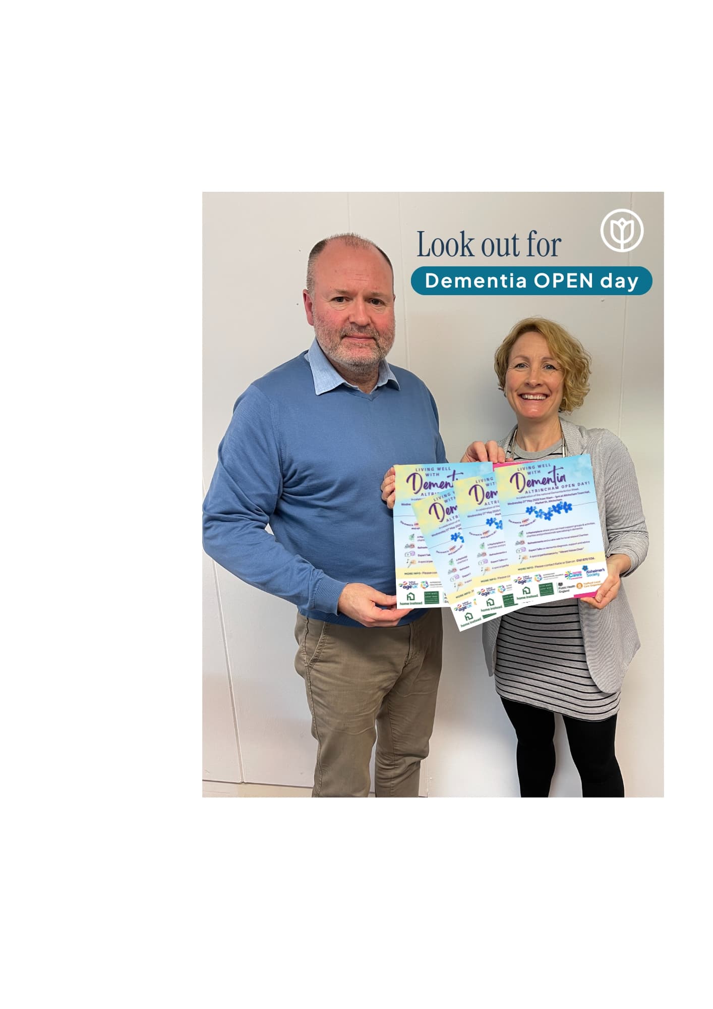 Two smiling people hold Dementia OPEN day flyers beside a sign that reads, "Look out for Dementia OPEN day. - Home Instead