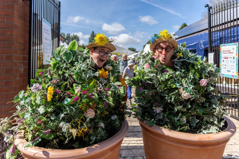 Two people in costumes resembling potted plants, with foliage and flowers, pose outdoors near an entrance gate. - Home Instead