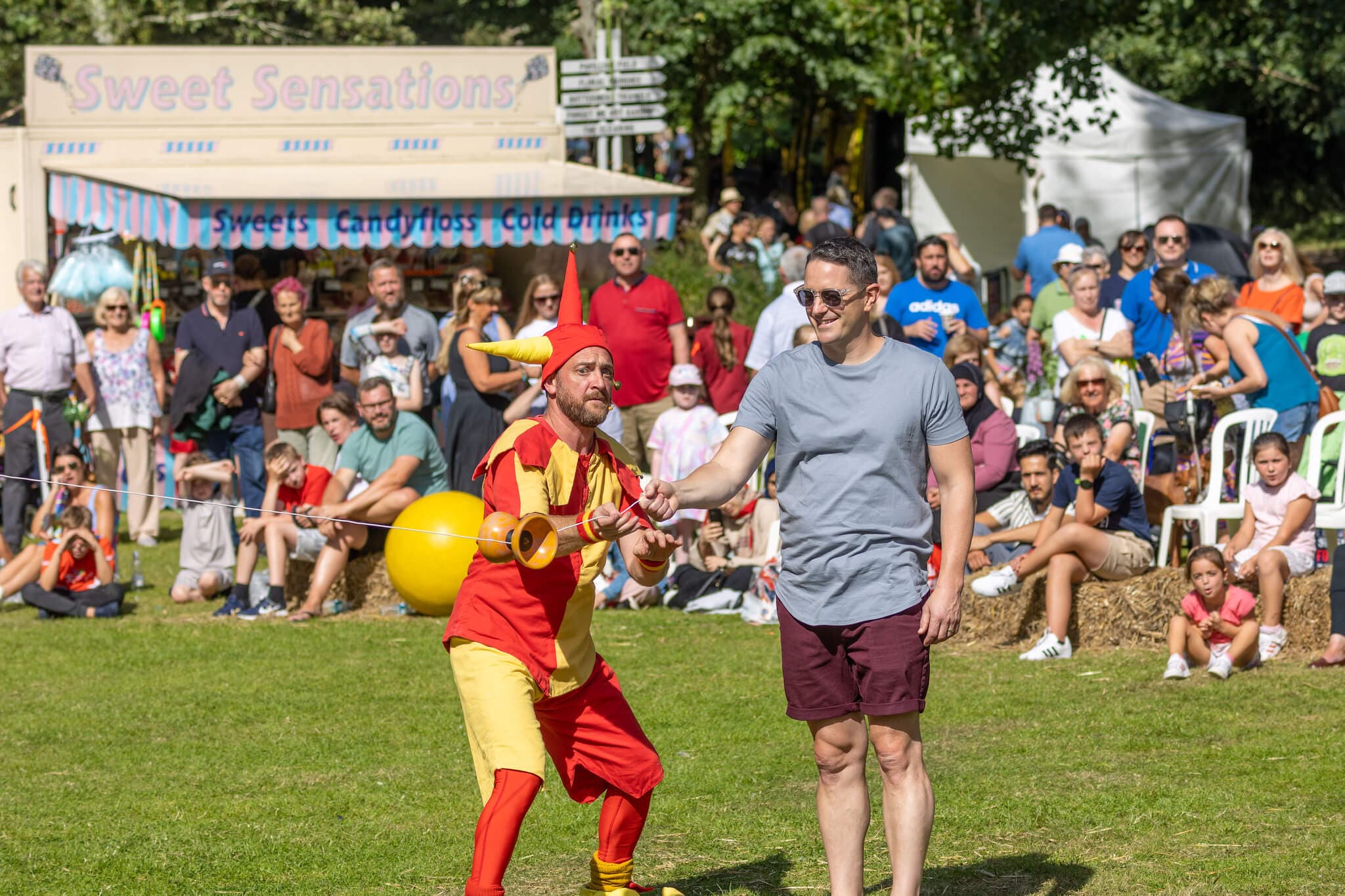 A jester performs with a balloon and a man at an outdoor event, with a crowd and food stall in the background. - Home Instead