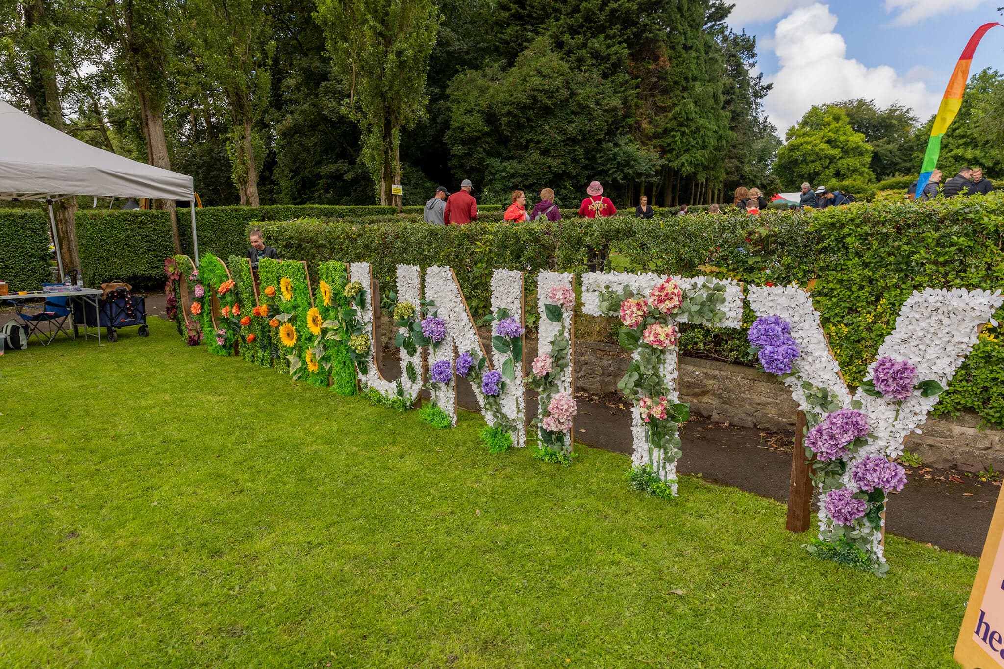 Floral letters spelling "COMMUNITY" on a grassy area with people and trees in the background. - Home Instead