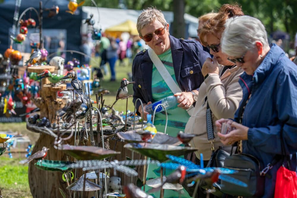 Three women browse colorful bird decorations at an outdoor market. Trees and vendor tents are visible in the background. - Home Instead