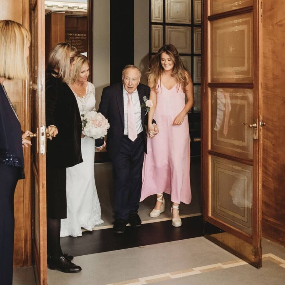 A bride in a white dress walks arm-in-arm with an elderly man, flanked by two women, and another woman in the foreground. - Home Instead