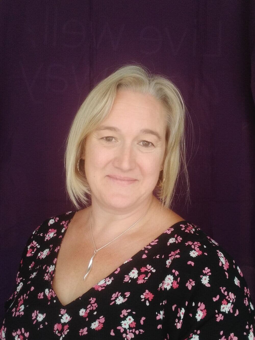 A blonde woman with a floral top smiles against a dark purple background. - Home Instead