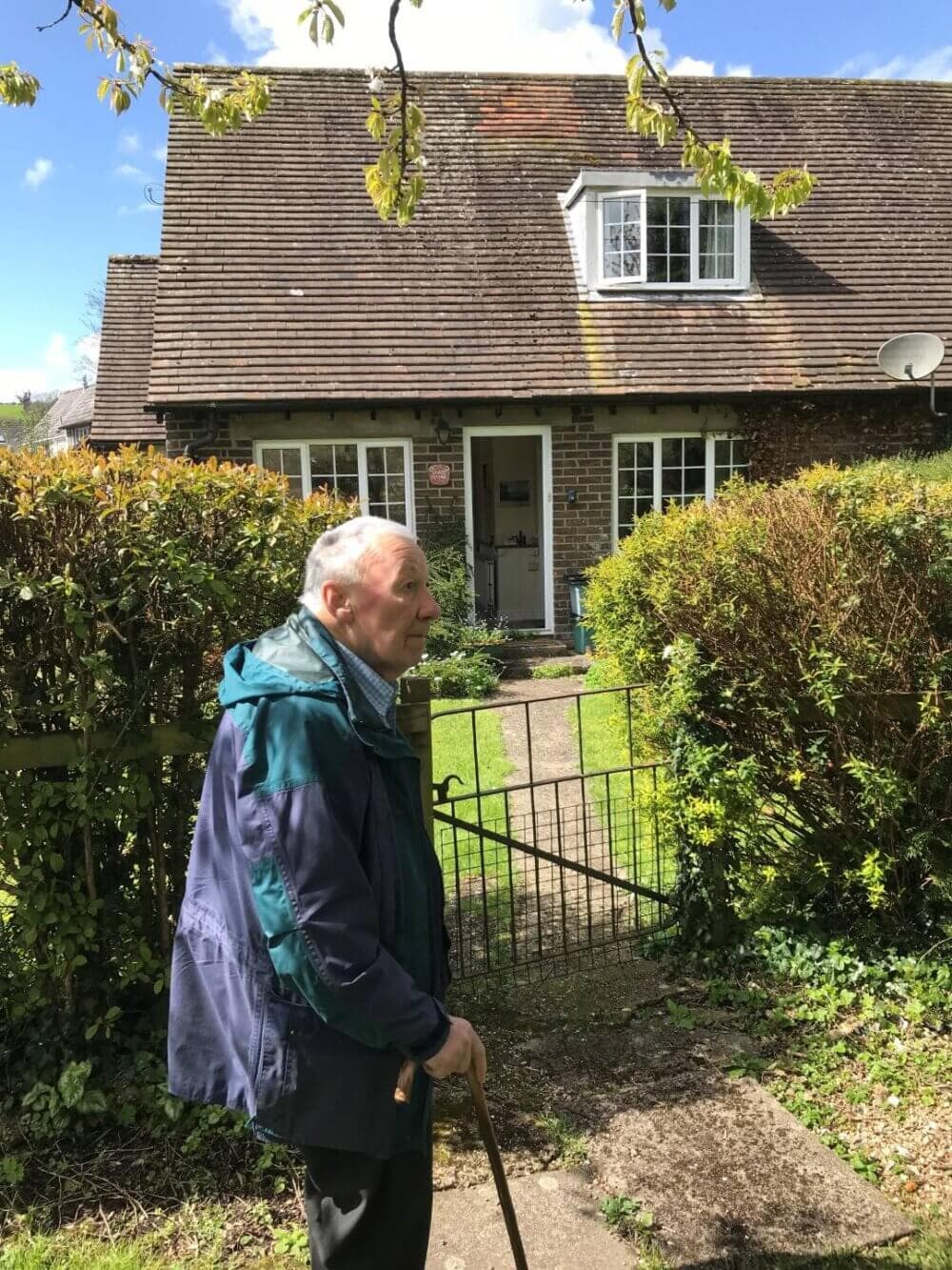 An elderly man with a cane stands in front of a brick house with a garden and hedge on a sunny day. - Home Instead