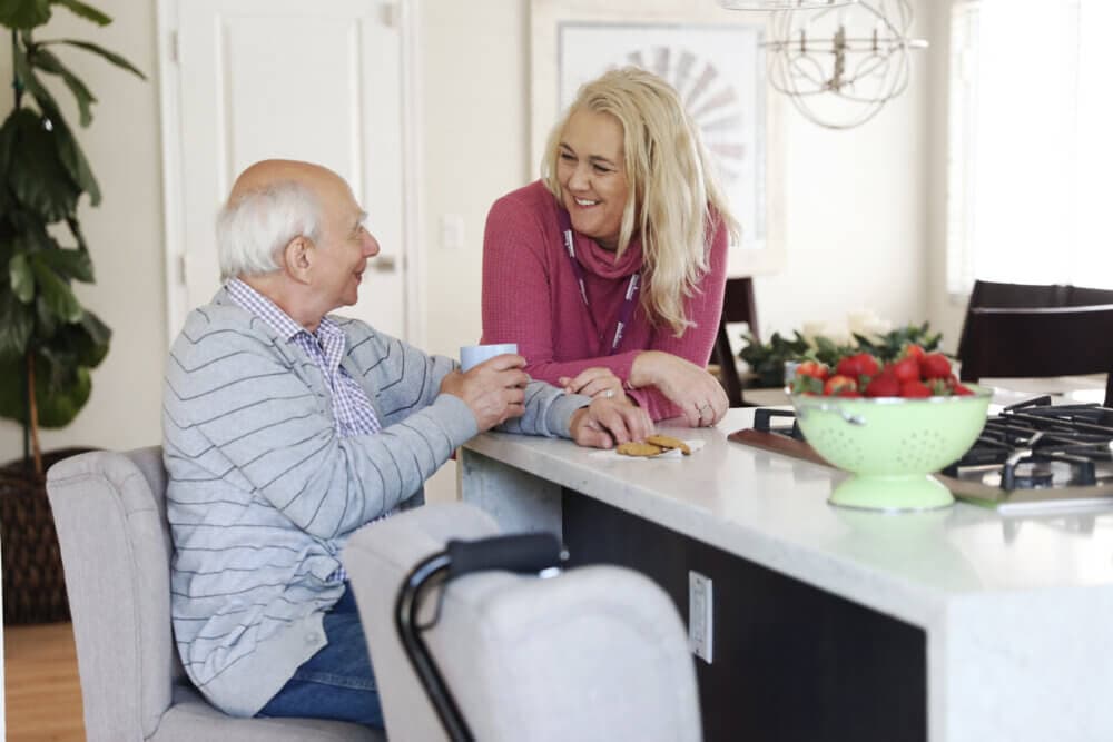 An elderly man and a woman share a conversation at a kitchen counter with a bowl of strawberries in the foreground. - Home Instead