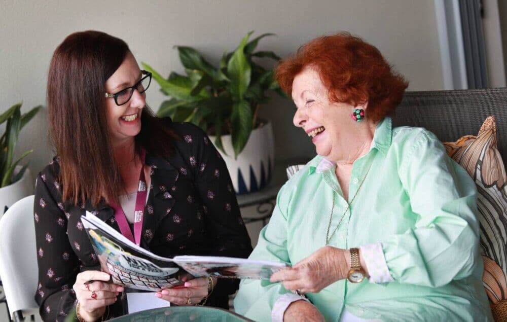 A woman and an elderly lady sit together, smiling and laughing while looking at a magazine. Plants are in the background. - Home Instead