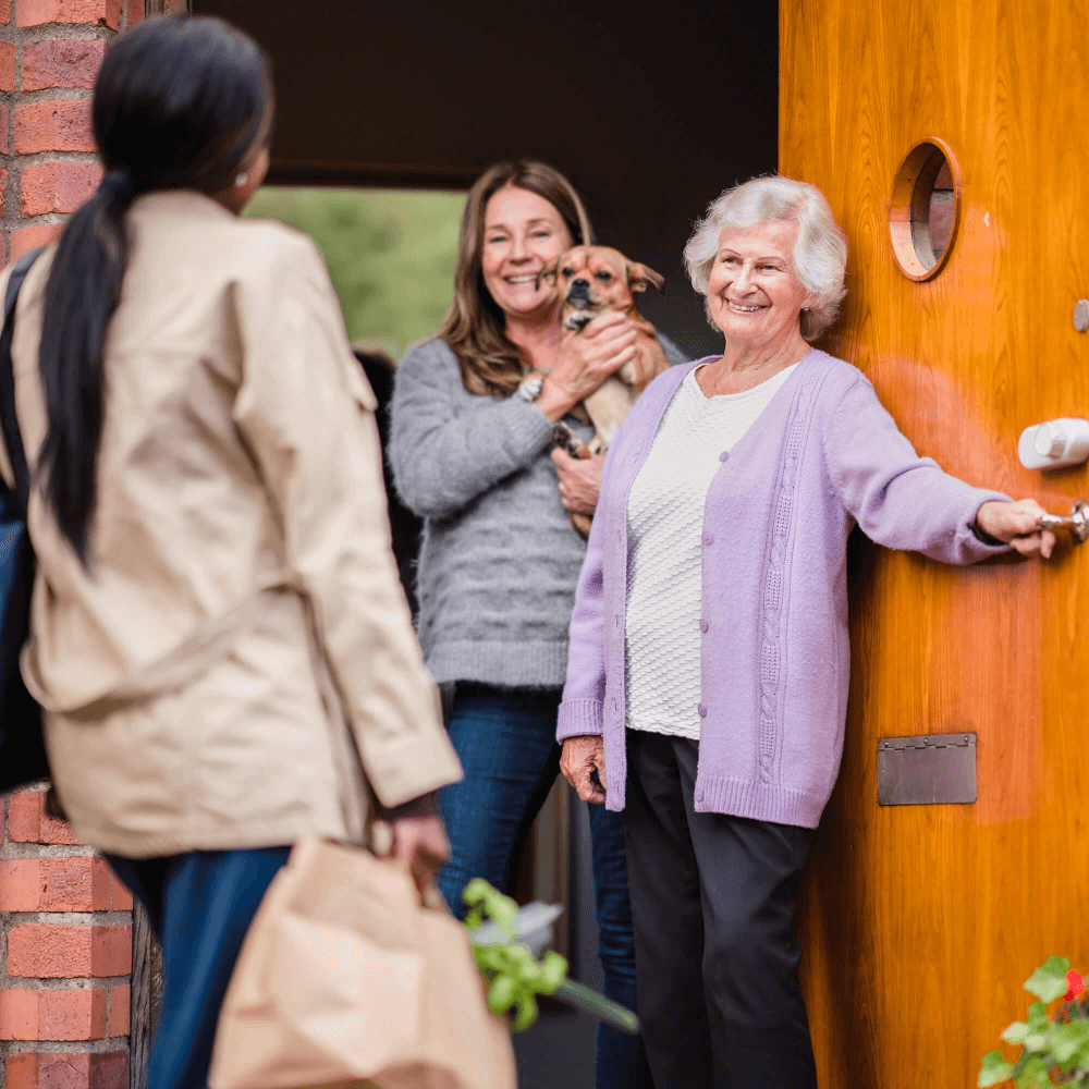 An elderly woman opens a door for a visitor holding groceries. A middle-aged woman holding a dog is in the background. - Home Instead