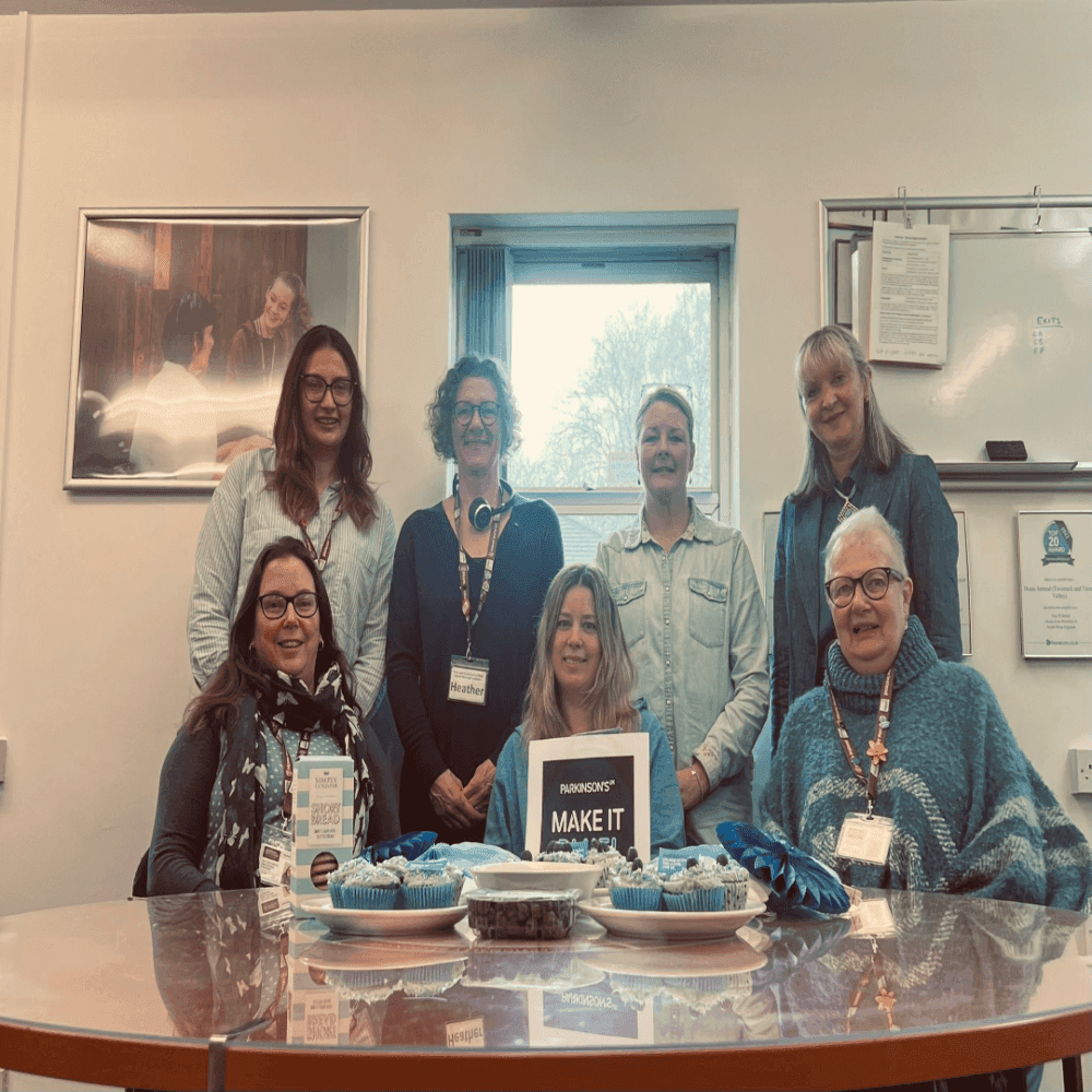 A group of seven people smiling around a table with baked goods and a sign that reads "MAKE IT. - Home Instead