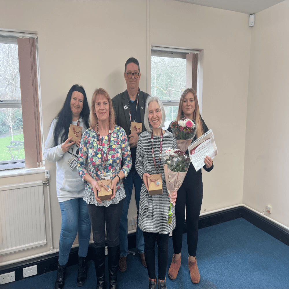 Five people are standing indoors, holding flowers and gifts, and smiling at the camera. - Home Instead