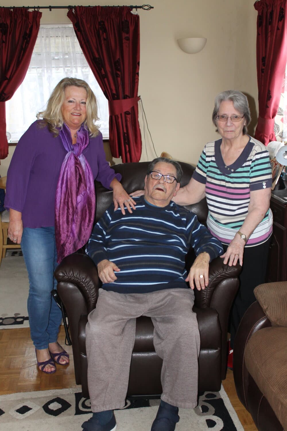 Three elderly individuals, two women standing beside a seated man in a home setting with red curtains. - Home Instead