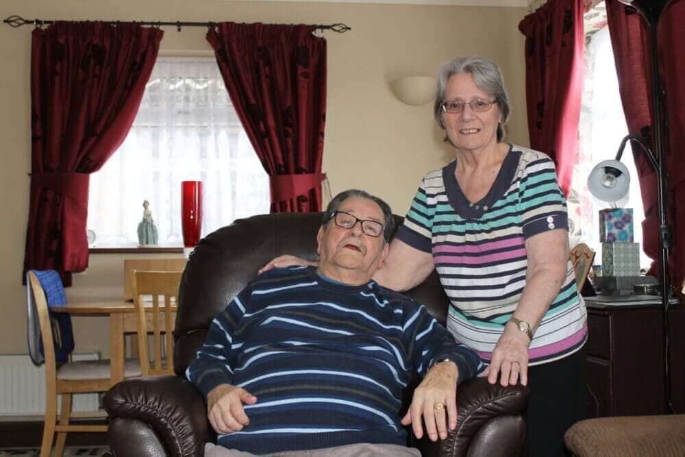 An elderly couple smiling, the man seated in a recliner and the woman standing beside him in a cozy living room. - Home Instead