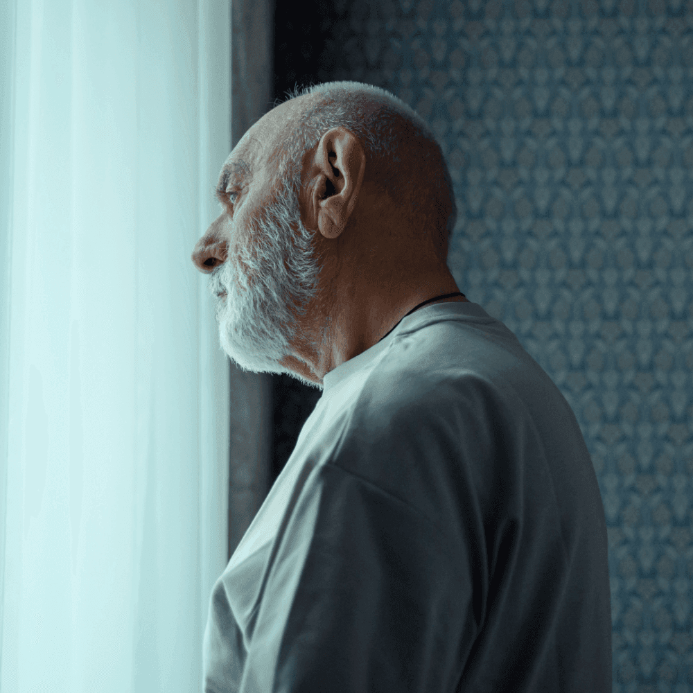 An elderly man with a beard stands by a window, looking outside as soft light illuminates his face. - Home Instead