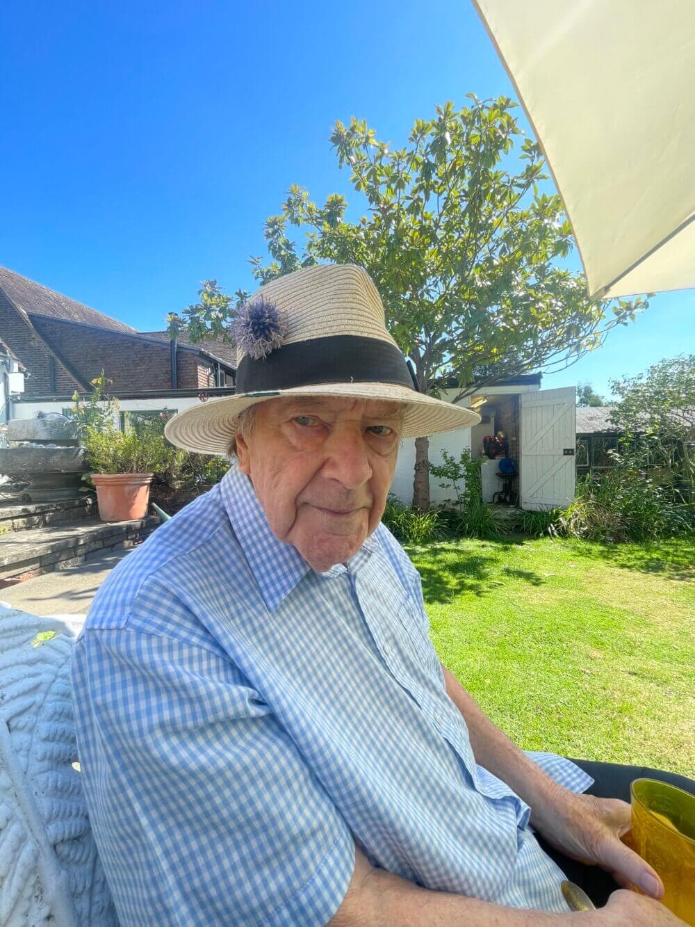 An elderly man wearing a hat sits outside under an umbrella, with a garden and shed in the background. - Home Instead