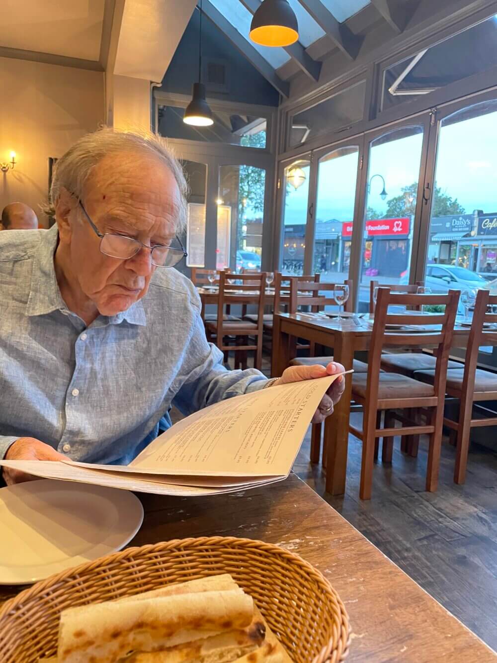 Elderly man with glasses reads a menu in a dimly lit restaurant with large windows. Basket of bread sits on the table. - Home Instead