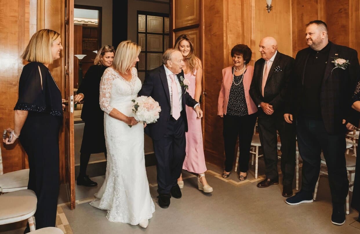 A bride in a white dress and a groom in a suit are celebrated by well-dressed guests in a warmly lit room. - Home Instead