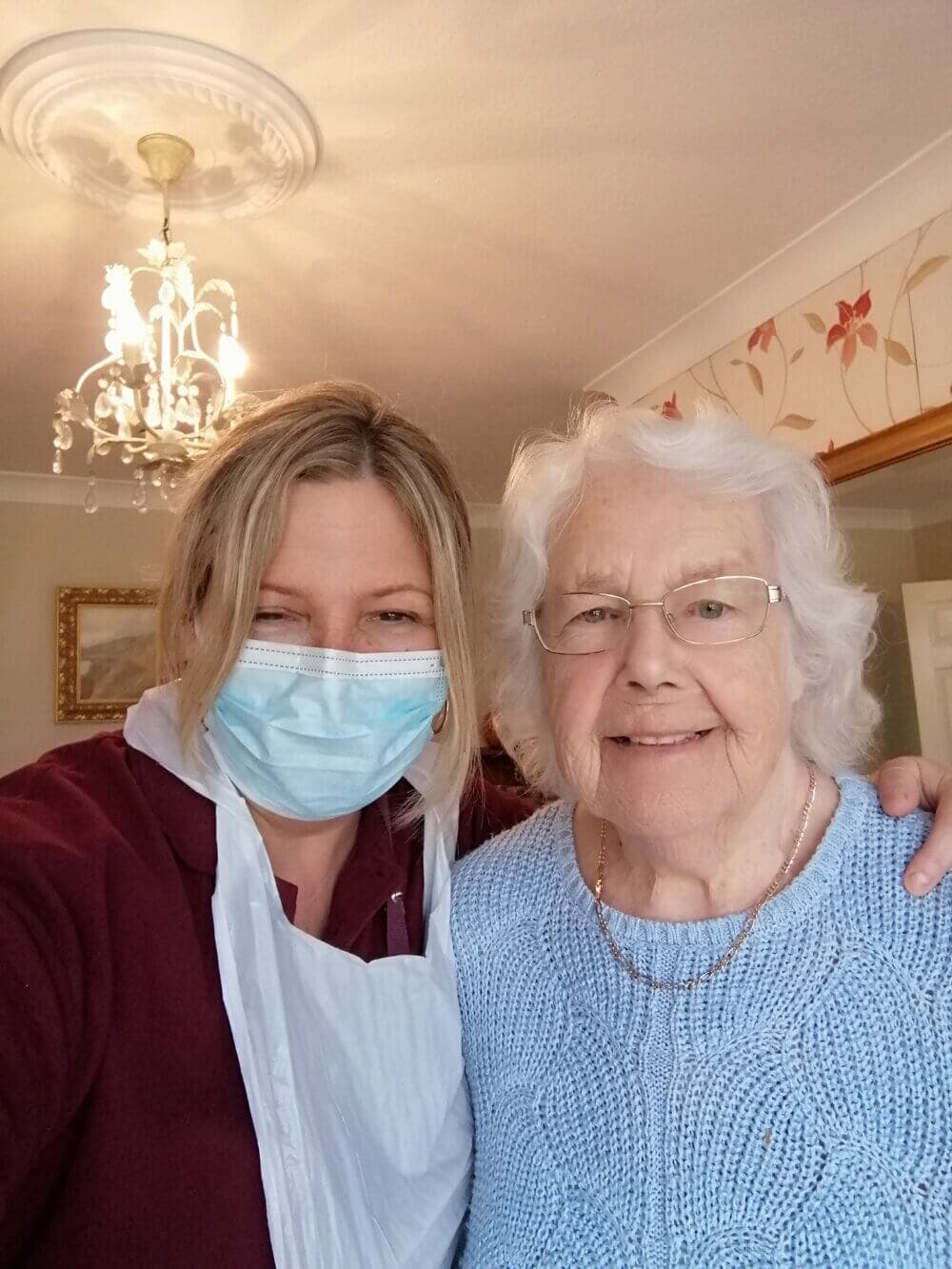 A younger woman in a mask taking a selfie with an older woman in glasses and a blue sweater, smiling indoors. - Home Instead