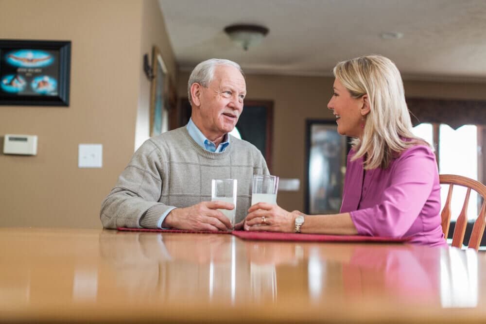 An elderly man and a middle-aged woman sit at a table, drinking milk and having a conversation. - Home Instead