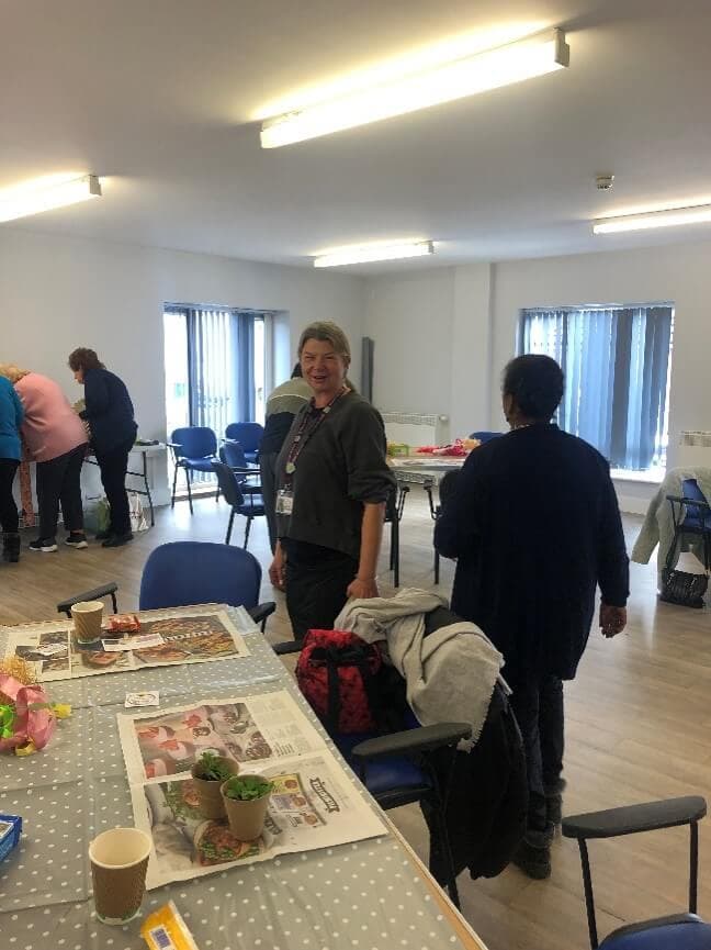 People in a room socializing and doing activities at tables, with newspapers and cups visible. - Home Instead