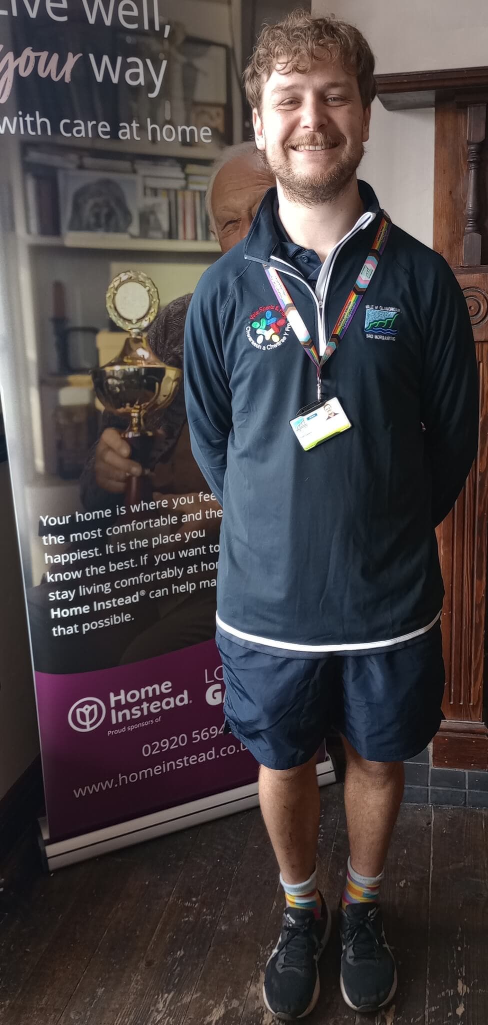A person wearing a uniform stands smiling in front of a Home Instead banner with ID badges and medals around their neck. - Home Instead