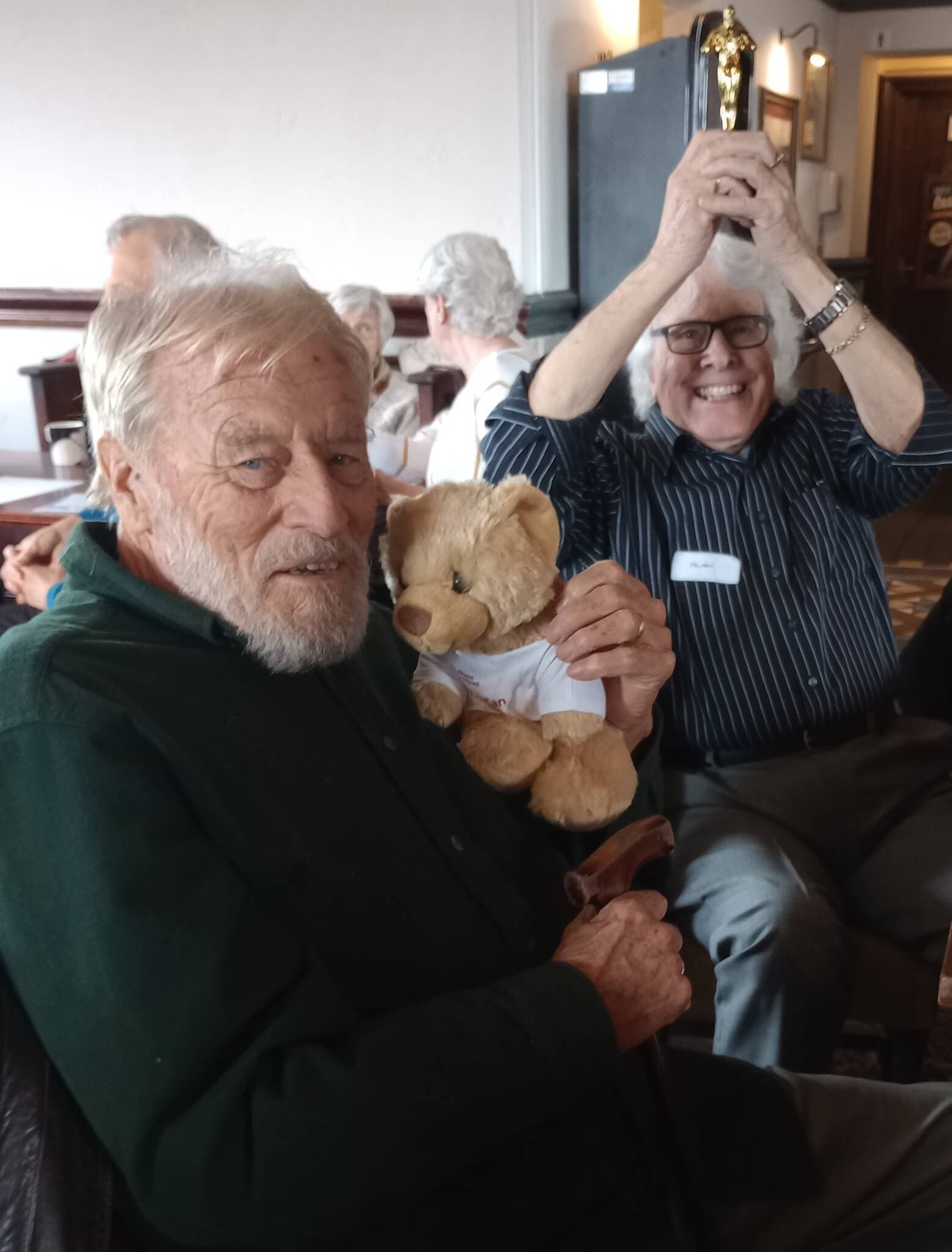 Two elderly men smiling; one holding a teddy bear, the other raising a trophy. People chatting in the background. - Home Instead