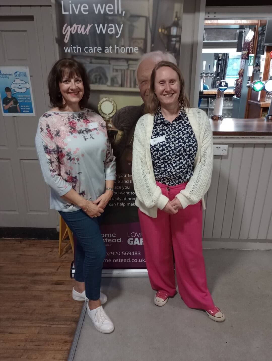 Two women standing and smiling in front of a banner with a text about home care services. - Home Instead