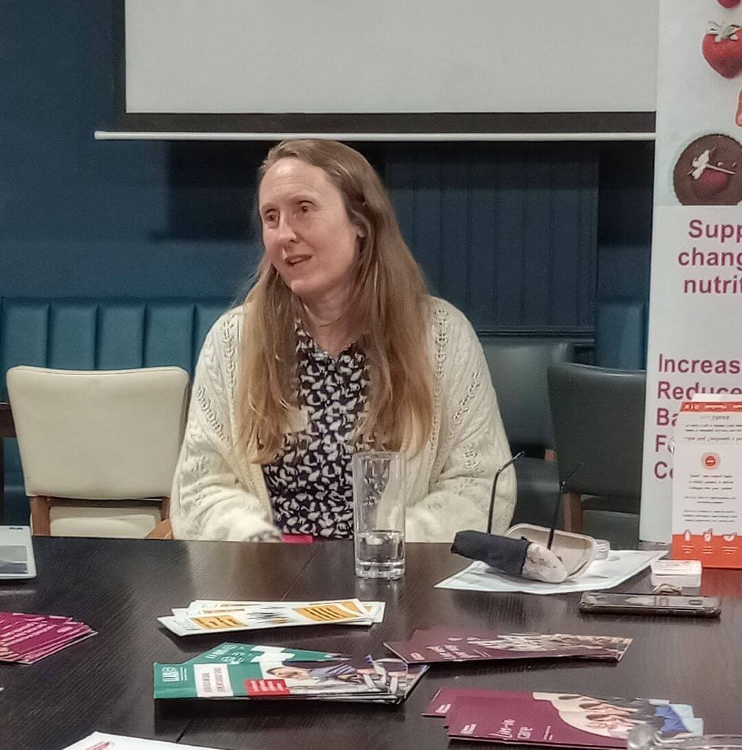 A person with long hair sits at a table with pamphlets and a glass of water, speaking in front of a banner. - Home Instead