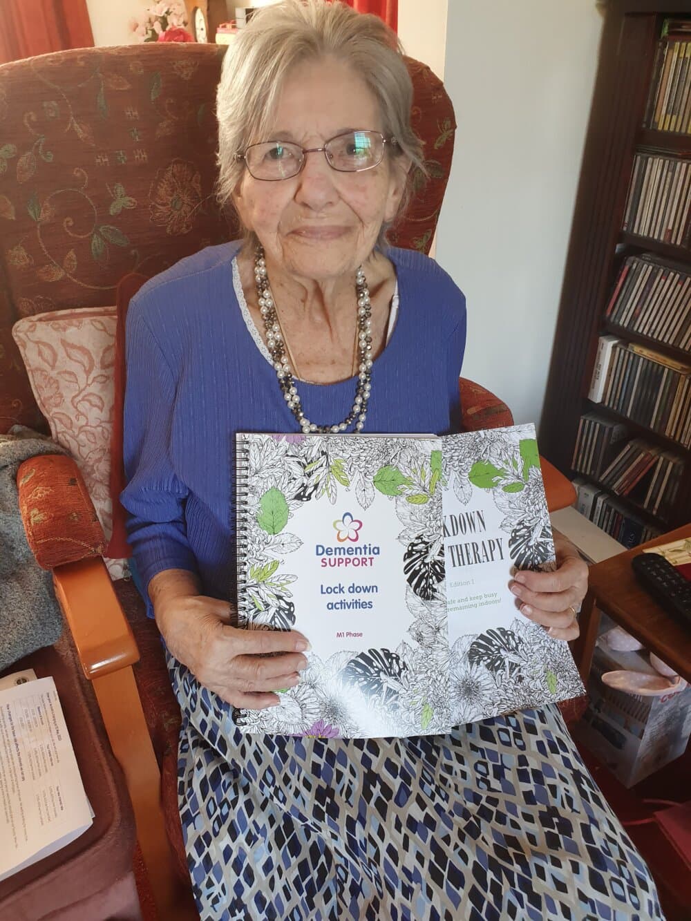 An elderly woman with glasses is sitting in a chair, holding a book titled "Lock down activities. - Home Instead