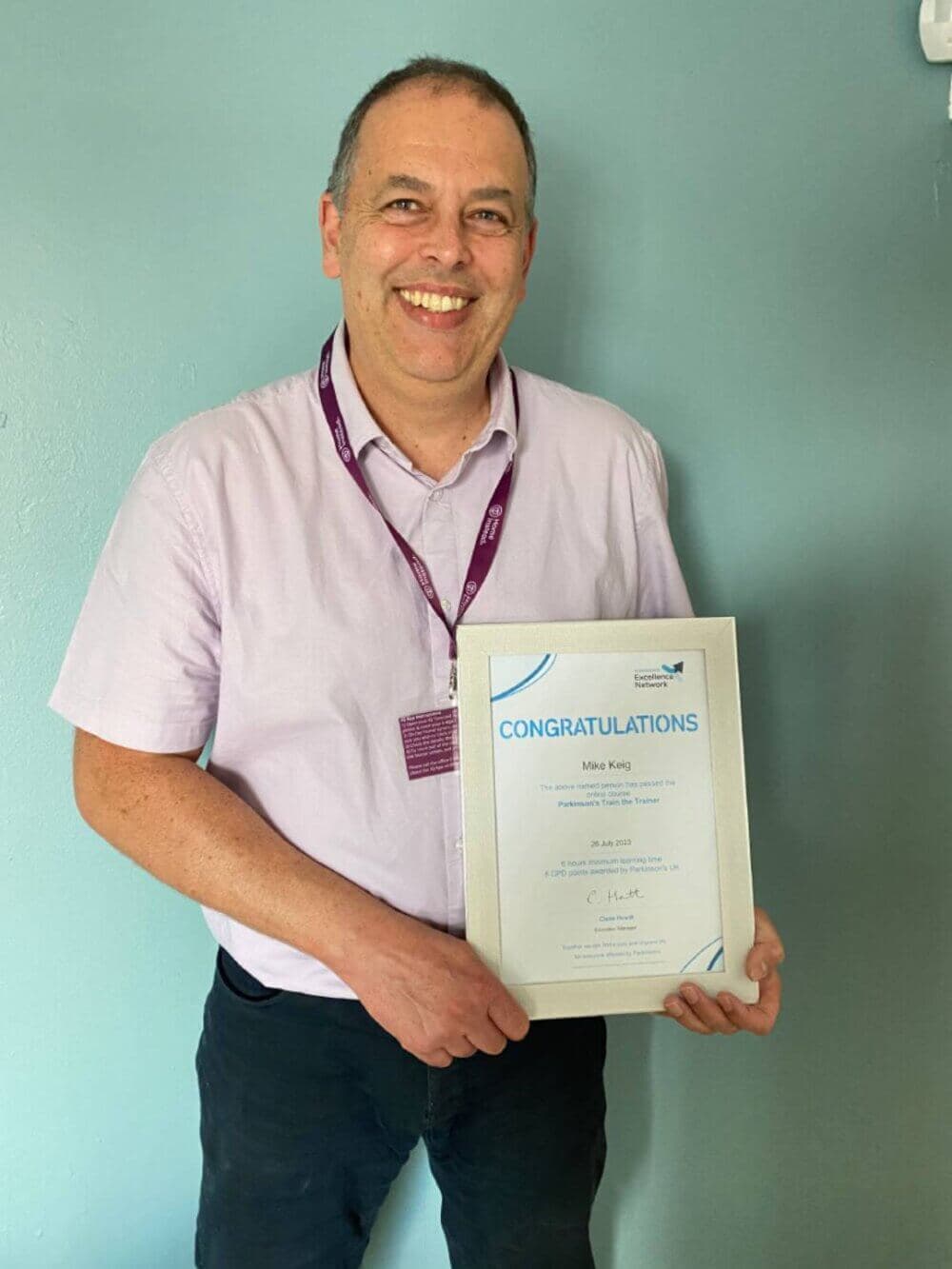 A man smiling and holding a certificate that reads "Congratulations." He is wearing a light purple shirt. - Home Instead