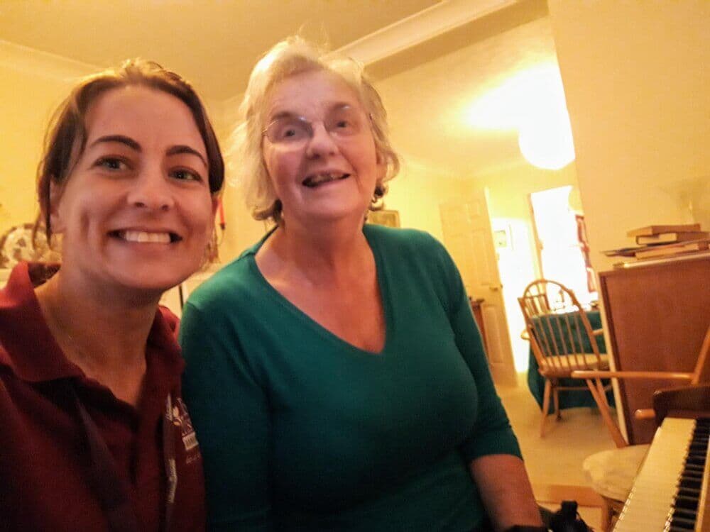 Two smiling women are sitting together indoors, one wearing a red shirt and the other wearing a green shirt. - Home Instead
