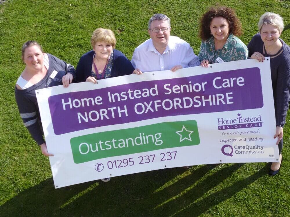 Five people holding a banner that reads "Home Instead Senior Care North Oxfordshire - Outstanding. - Home Instead