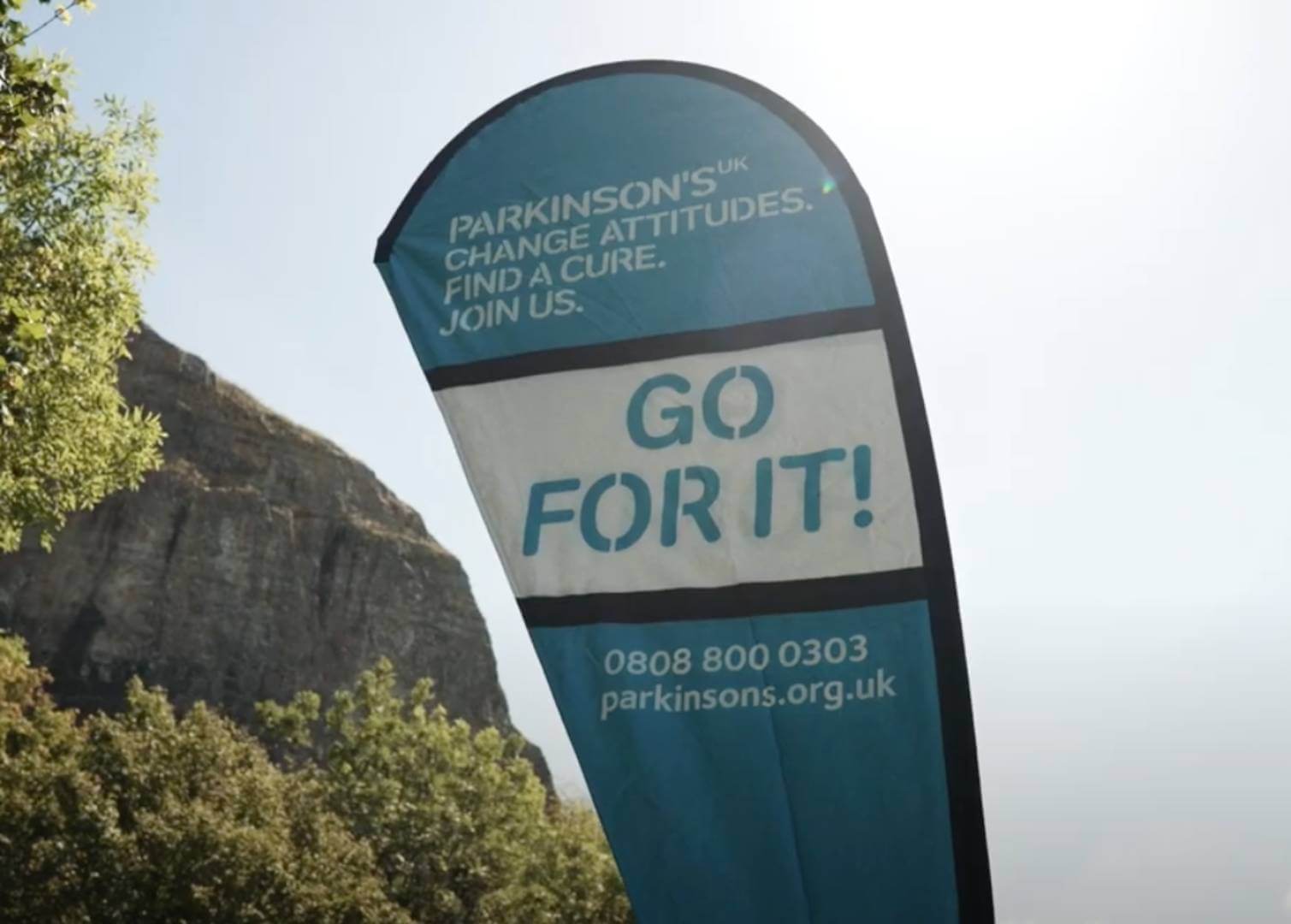 Blue and white Parkinson's UK banner with the slogan "GO FOR IT!" and contact details. Trees and rock in the background. - Home Instead