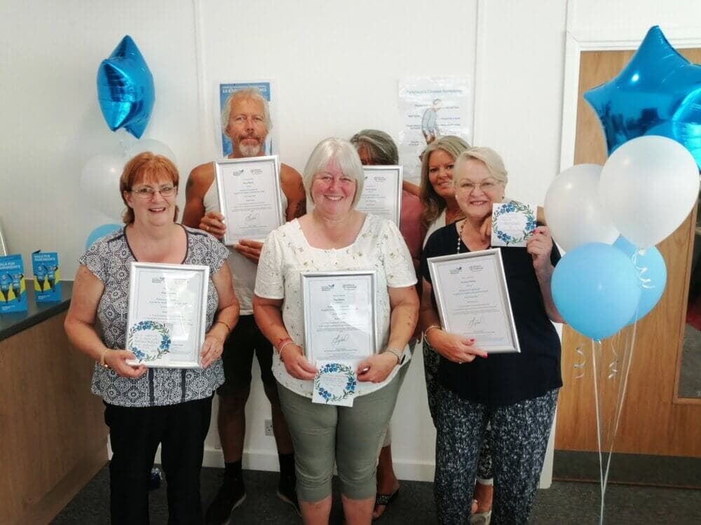 Six people holding certificates and balloons, celebrating in a decorated room with white and blue balloons. - Home Instead