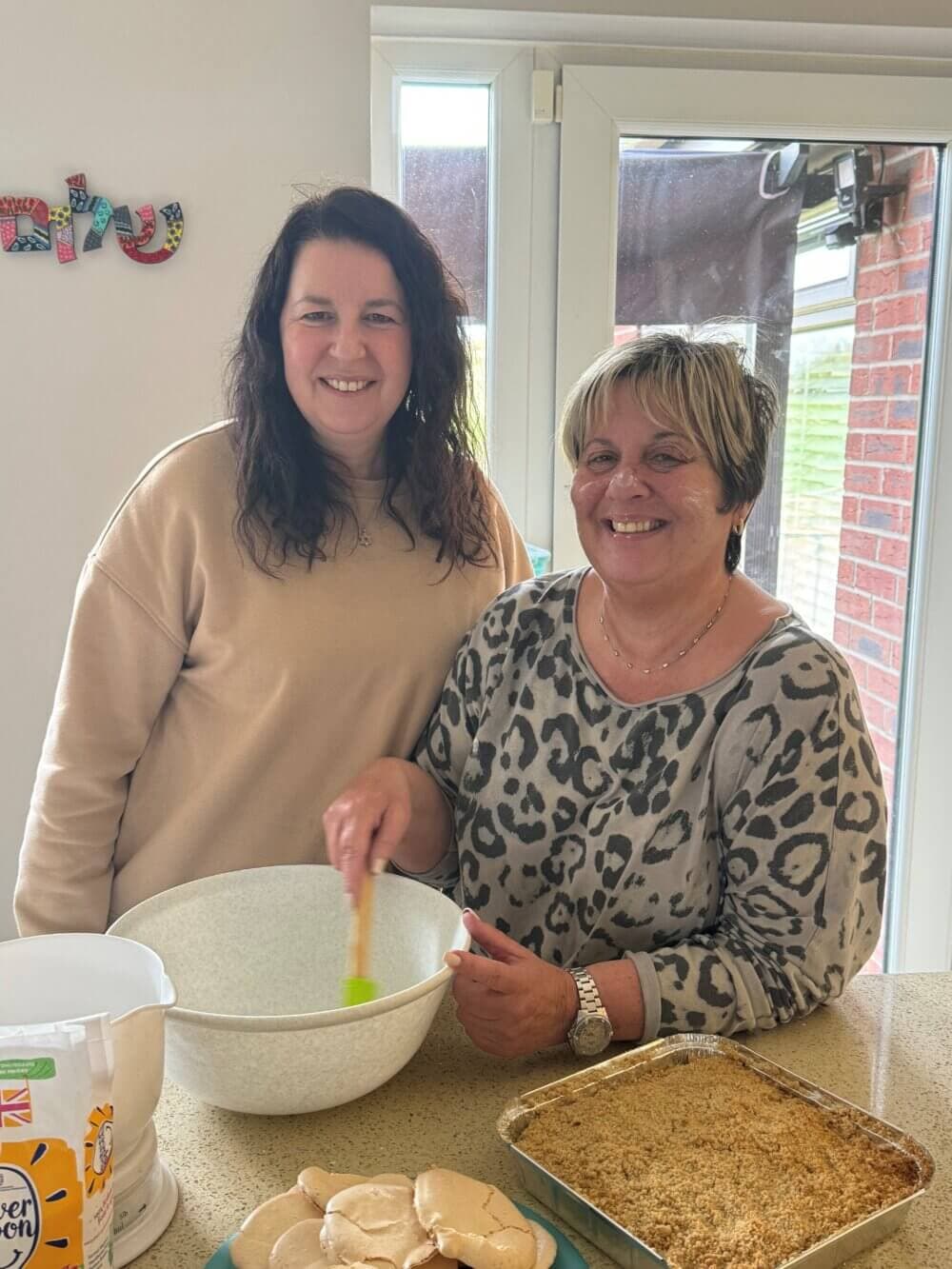 Two smiling women baking together in a kitchen, one stirring a bowl while the other poses beside her. - Home Instead