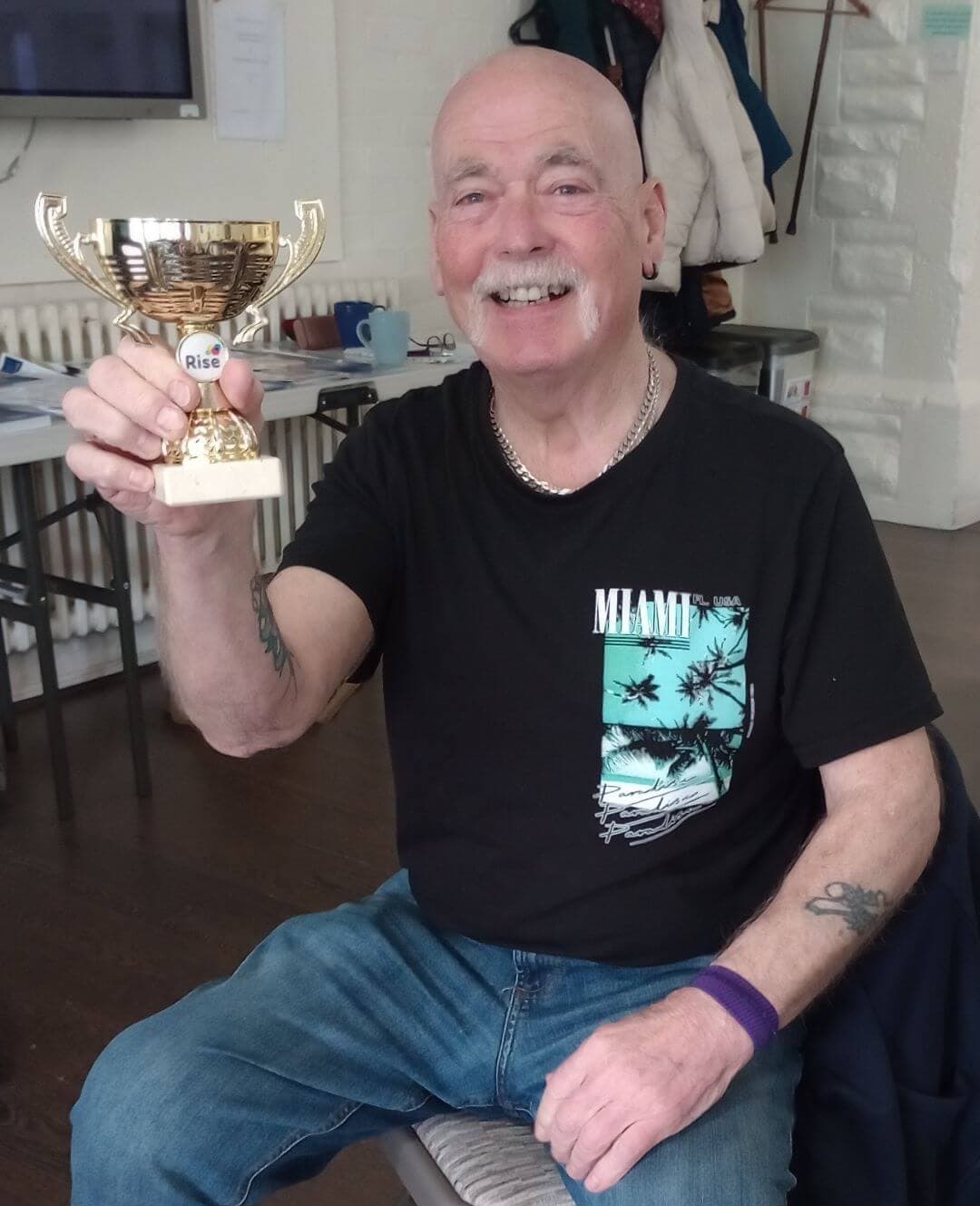Smiling man with a moustache holding a gold trophy while sitting, wearing a black shirt and blue jeans in a room. - Home Instead