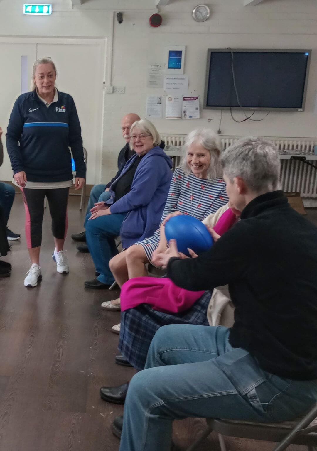 A group of elderly people sits in a circle passing a blue ball to each other in a community center. - Home Instead