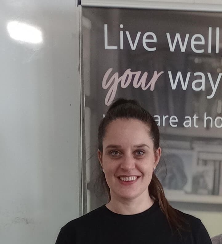 Smiling woman with a ponytail standing in front of a whiteboard with the text "Live well your way. - Home Instead