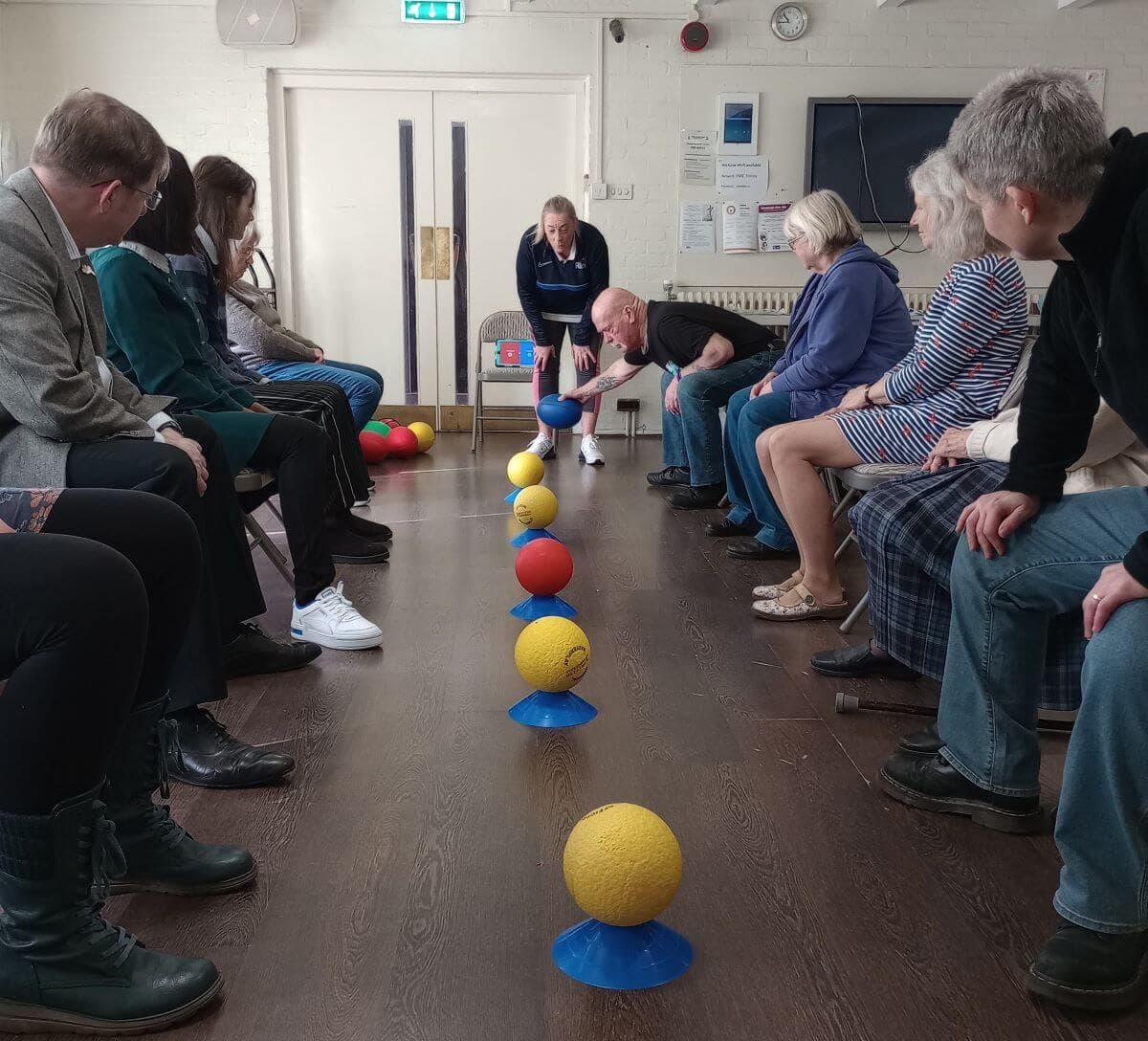 A group of people sits in a semicircle, watching a man roll a ball towards other colored balls lined up on the floor. - Home Instead
