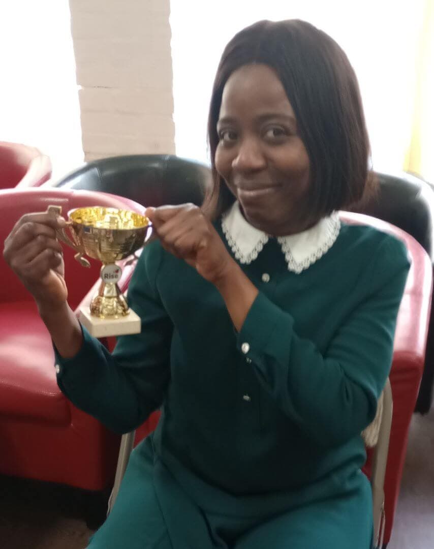 A woman holds up a trophy while smiling, seated on a chair in front of a red couch. - Home Instead