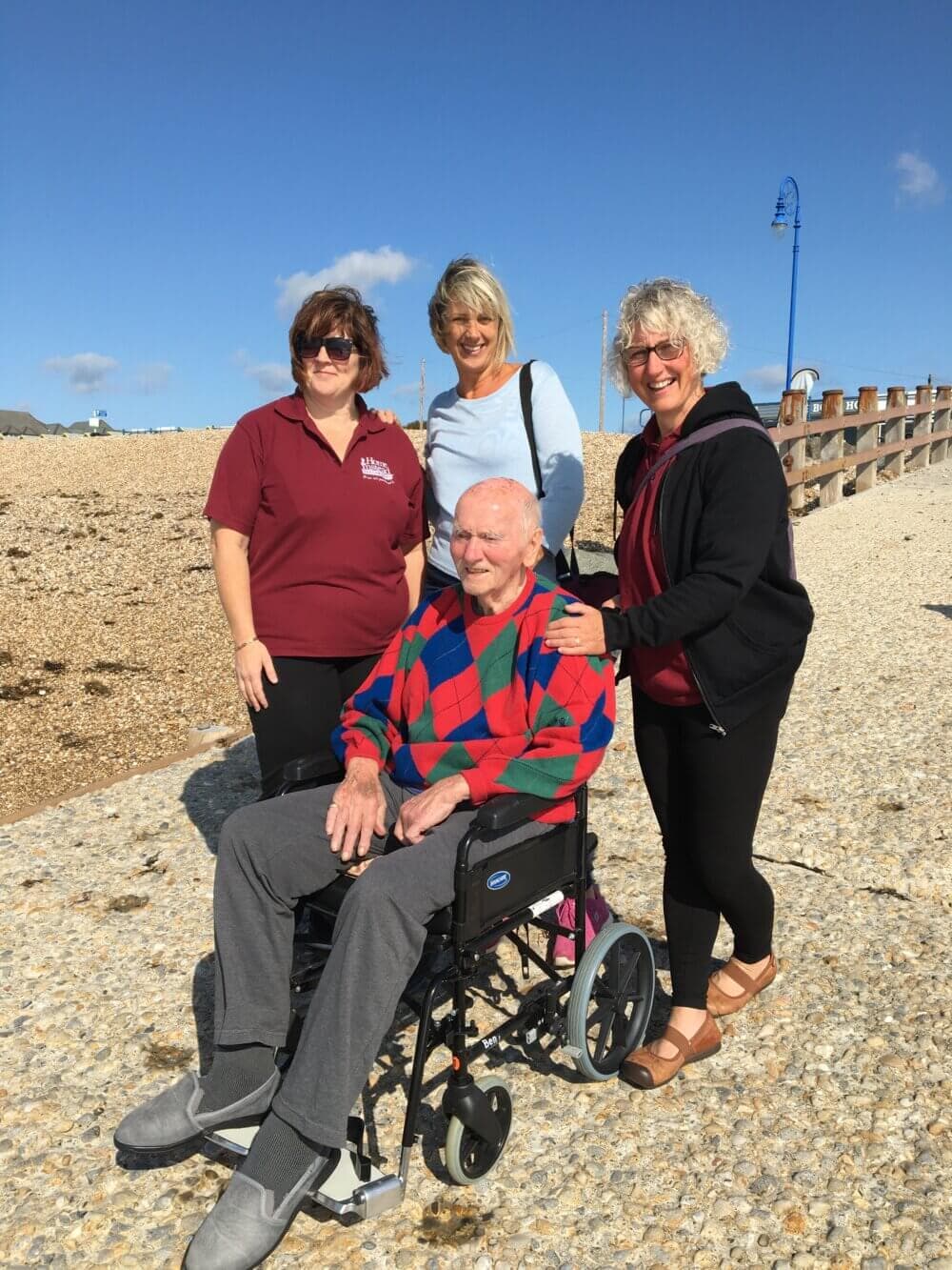 Four people smiling on a sunny beach; one person is seated in a wheelchair, and the others are standing around them. - Home Instead