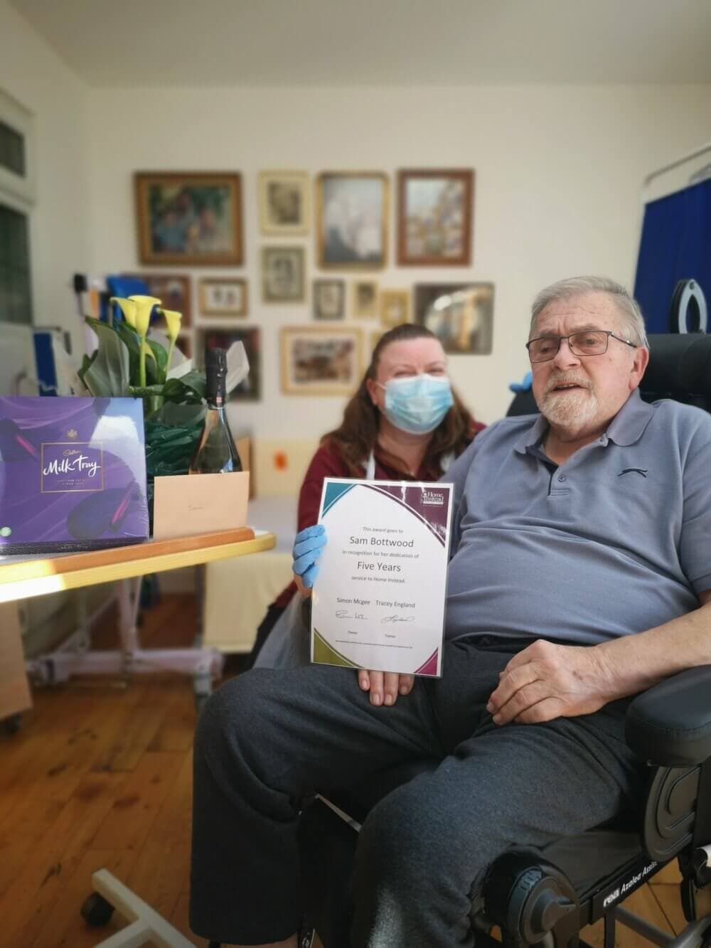 Man in a wheelchair holding a certificate with a woman standing beside him, holding a bouquet and a gift box. - Home Instead