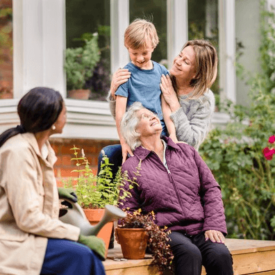 A young boy, his mother, grandmother, and another woman smile together on a bench in a garden. - Home Instead