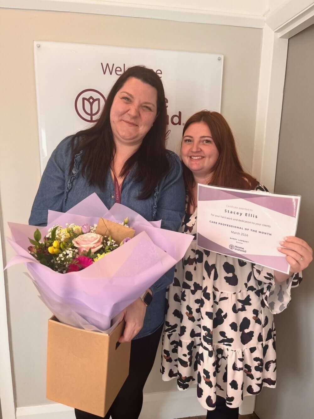 Two women smiling, one holding a bouquet of flowers and the other holding a certificate labeled "Stacey Ellis. - Home Instead