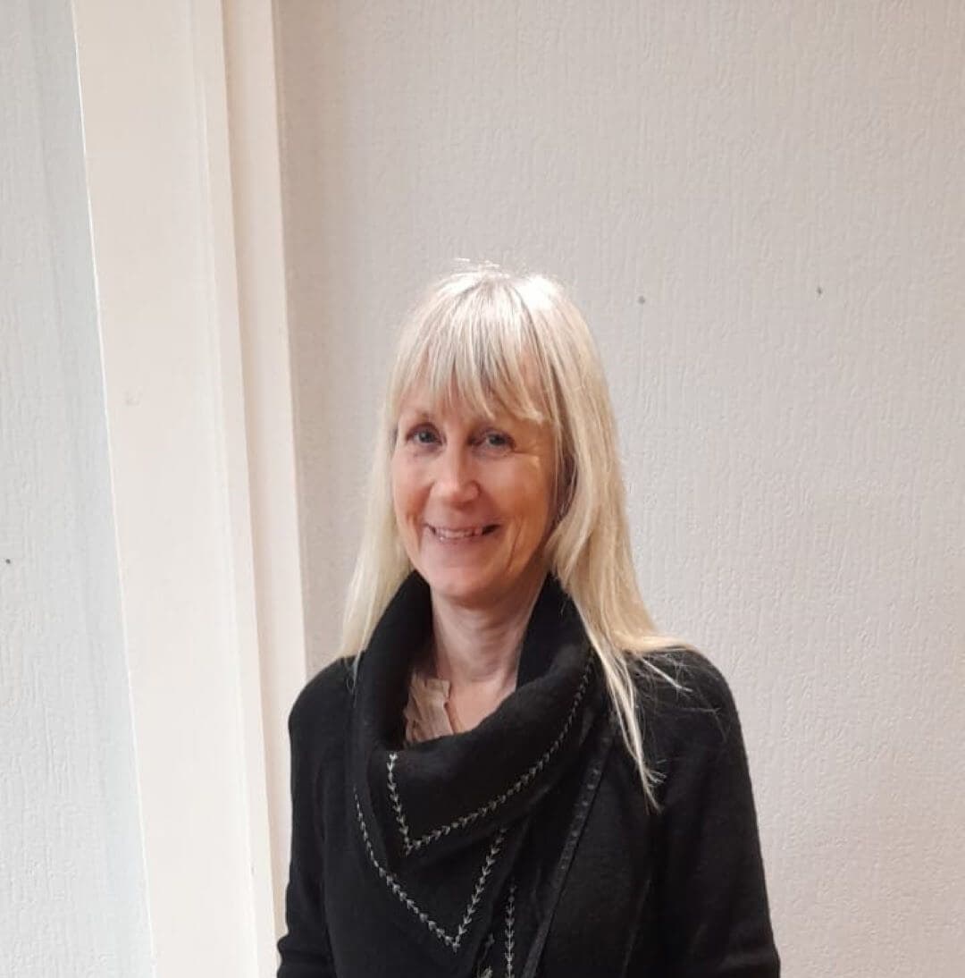Smiling person with long blonde hair, wearing a black top, stands against a light-colored wall. - Home Instead