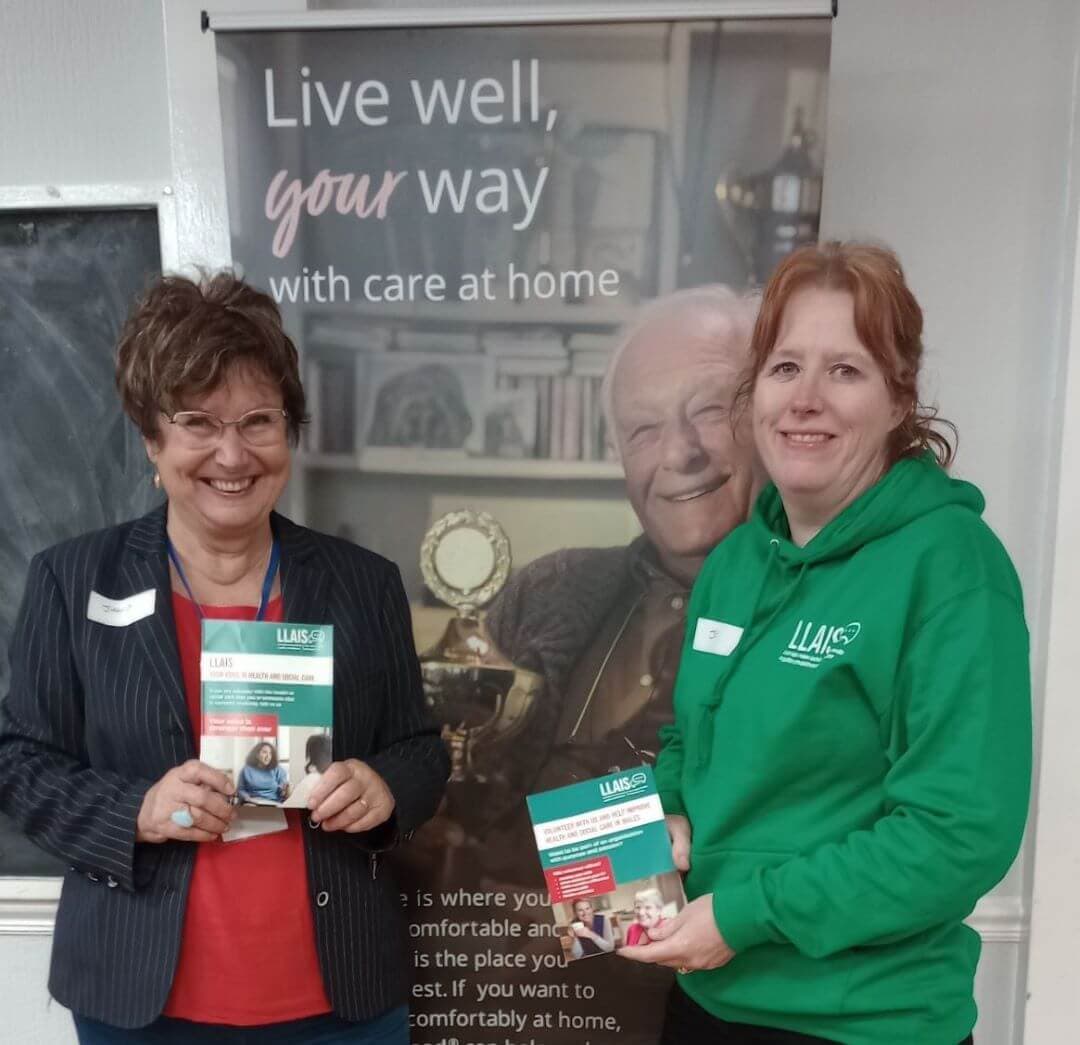 Two smiling women holding pamphlets by a sign that reads "Live well, your way with care at home" with an elderly man. - Home Instead