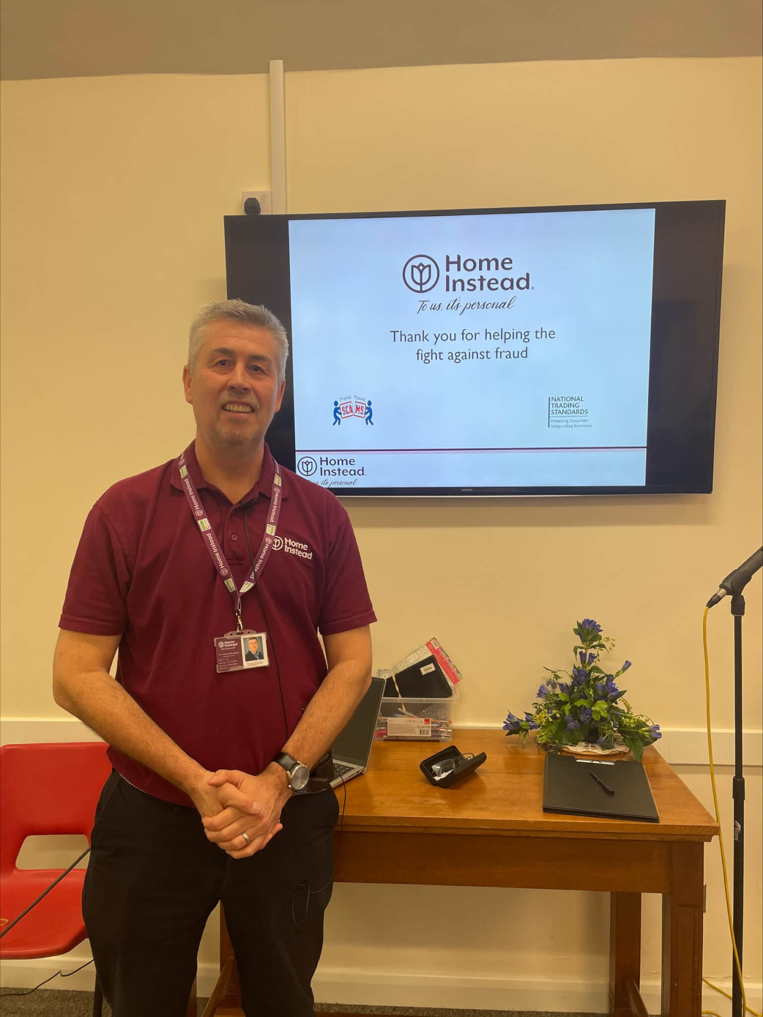 A man standing in front of a screen displaying "Home Instead" text and anti-fraud message in a room with a red chair. - Home Instead