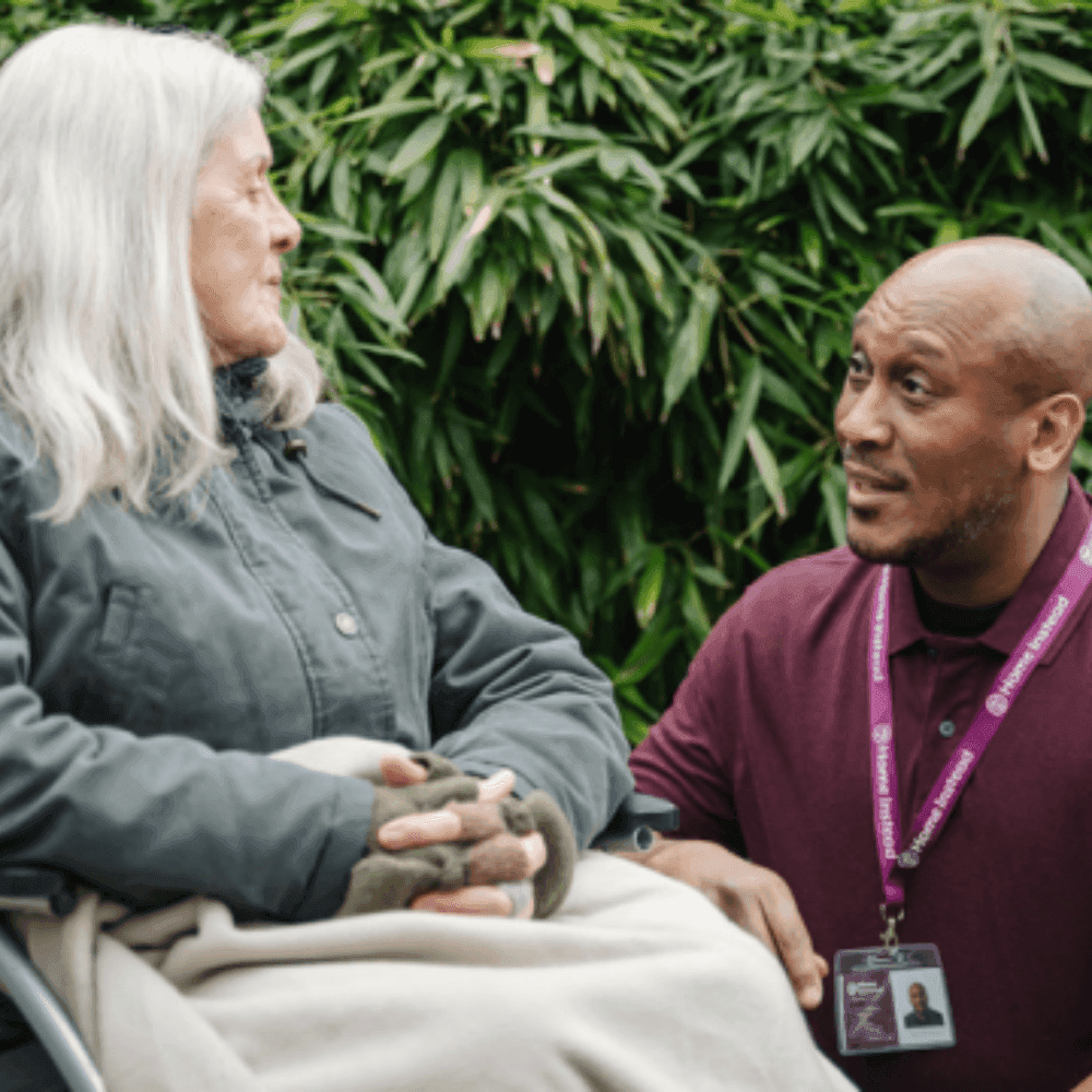 An older woman in a wheelchair converses with a man wearing a burgundy shirt and a lanyard in a garden. - Home Instead
