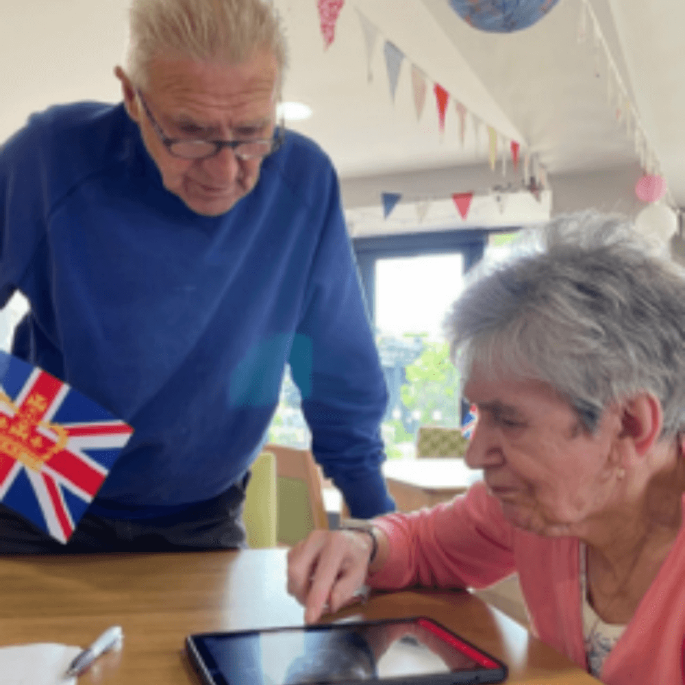 Two elderly individuals, one holding a flag, look at a tablet together on a table in a decorated room. - Home Instead