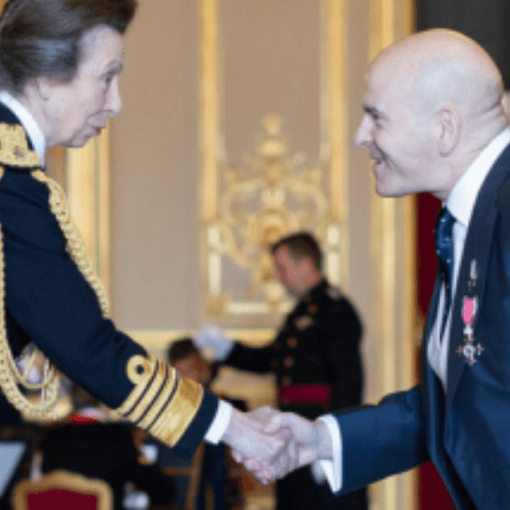 A woman in military uniform shakes hands with a man in a suit, both smiling, in a formal setting. - Home Instead