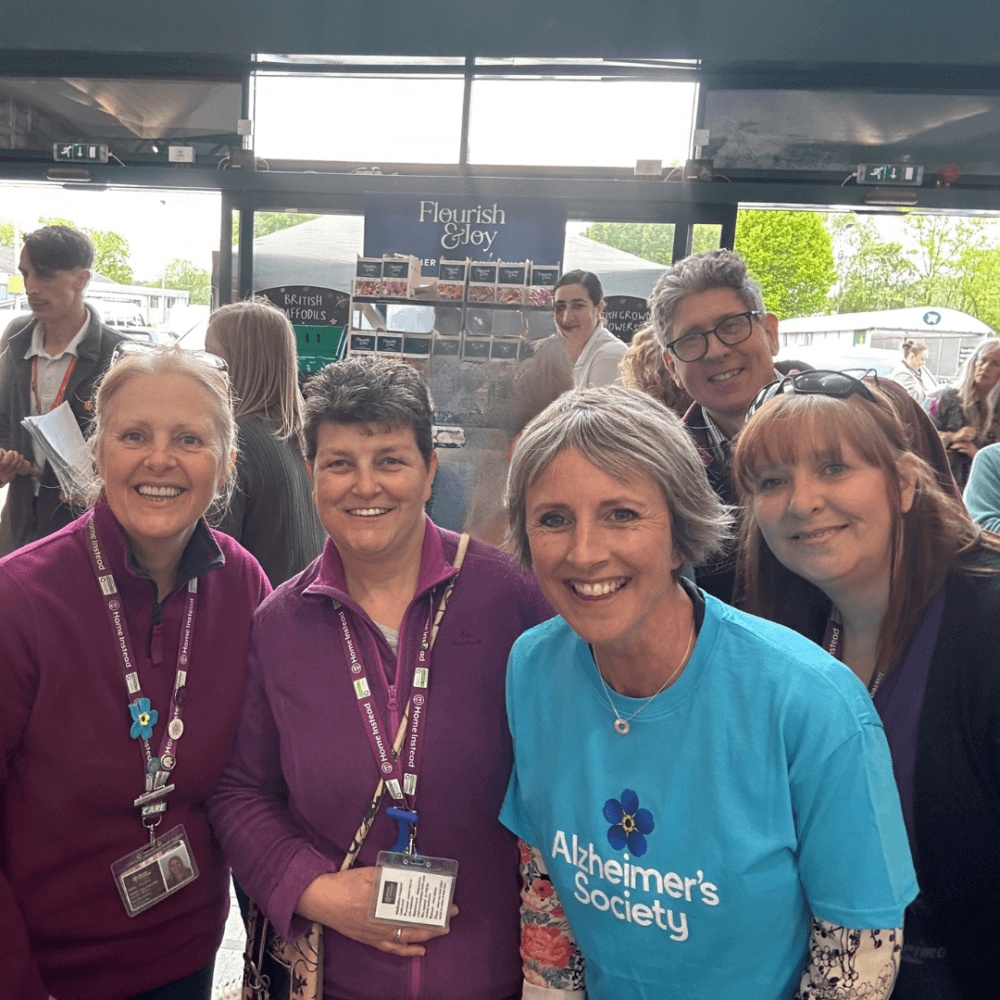 A group of people, smiling, gather indoors; one person wears an "Alzheimer's Society" shirt, others wear purple. - Home Instead