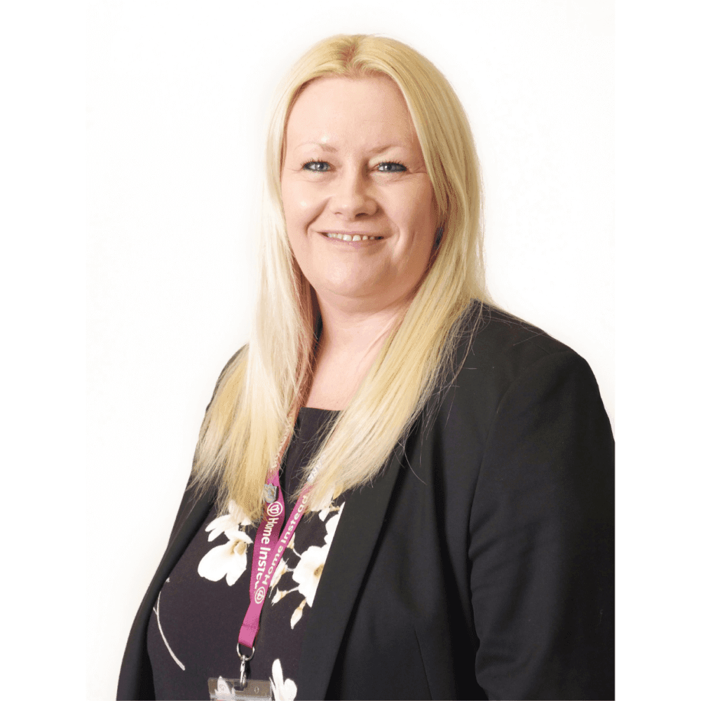 Smiling person with long blonde hair wearing a black blazer and a lanyard, standing against a white background. - Home Instead